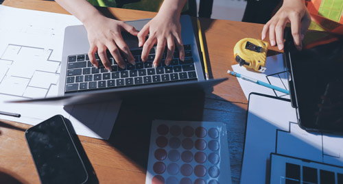 Hands typing on a laptop surrounded by blueprints, a tape measure, and a smartphone on a wooden table.